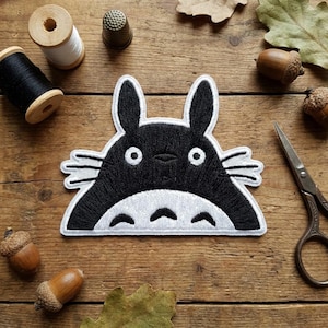 May include: Black and white embroidered patch featuring a cartoon character with large ears and a round body. The patch is surrounded by sewing supplies, acorns, and leaves on a wooden surface.