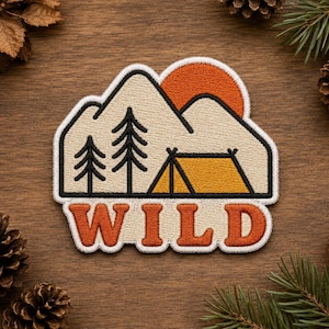 May include: An embroidered patch featuring a beige mountain landscape with two black pine trees, a yellow tent, and a burnt orange sun. The word WILD is stitched in large, bold, burnt orange letters at the bottom of the patch with a white border.
