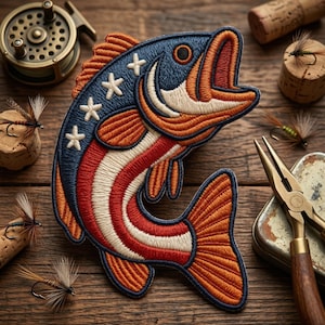May include: Embroidered patch in the shape of a fish, featuring a patriotic design with red and white stripes, blue with white stars, and orange accents. Fishing-themed items, including a reel, flies, and pliers, surround the patch on a wooden surface.