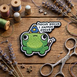 May include: Embroidered patch featuring a green frog wearing a wizard hat and holding a wand. The patch has the text "hocus pocus I cannot focus". The patch is surrounded by sewing supplies and lavender sprigs.