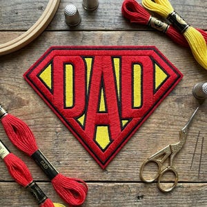 May include: A red and yellow embroidered patch in a diamond shape with the word "DAD" in black. The patch is surrounded by red embroidery floss, yellow and red thread spools, thimbles, scissors, and needles on a wooden surface.