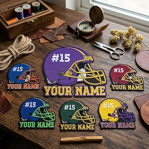 May include: A collection of embroidered football helmet patches in various colors, including purple, blue, maroon, green, and yellow. Each patch features the number "15" and the text "YOUR NAME" in a contrasting color. The patches are displayed on a wooden surface.