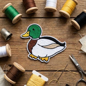 May include: A colorful embroidered duck patch with a green head, brown chest, and white body. The patch is surrounded by spools of thread in various colors, a thimble, scissors, and a needle on a wooden surface. Crafting and sewing supplies.