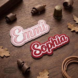 May include: Emmi and Sophia embroidered name patches in a script font with a white border. The Emmi patch is pink, and the Sophia patch is red. The patches are on a wooden surface with decorative elements.