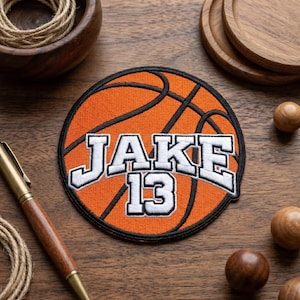 May include: An orange basketball patch with black lines and the name "JAKE" and the number "13" in white, outlined in black. The patch is round and sits on a wooden surface.