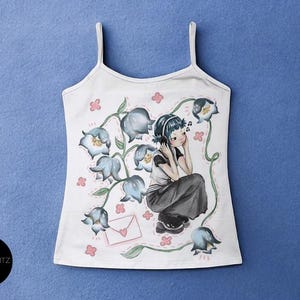 May include: White tank top with a floral illustration featuring a girl wearing headphones, surrounded by blue flowers and pink accents. The design includes a letter with a heart. The brand name "HYPERGLITZ" is on the bottom left.
