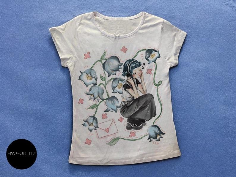 May include: White t-shirt featuring a seated anime-style character wearing headphones, surrounded by bluebell flowers and pink accents. The design includes a letter with a heart. The shirt has short sleeves and a round neckline.