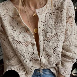 Puede incluir: Un cárdigan de crochet beige con botones y mangas largas. El diseño de tejido abierto revela la ropa de debajo. El cárdigan se combina con vaqueros azules y collares dorados. La palabra "HYPERGLITZ" es visible en la imagen.