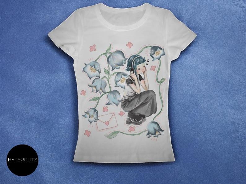 May include: White t-shirt featuring a watercolor illustration of a person wearing headphones, surrounded by blue flowers and musical notes. A pink envelope is also in the design. The shirt has a crew neck and short sleeves.