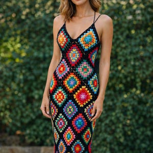 Może przedstawiać: Kolorowa, ręcznie robiona sukienka z szydełka z dekoltem w kształcie litery V i cienkimi ramiączkami. Sukienka ma czarne tło z żywymi, kwadratowymi wzorami w odcieniach czerwieni, pomarańczy, żółci, zieleni, niebieskiego i różu. Sukienka jest dopasowana i długa.