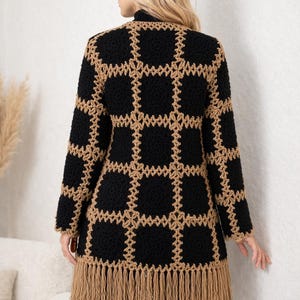 Op de afbeelding: Lange, open vest in zwart en beige met een ruitpatroon. Het haakontwerp heeft een raster van zwarte vierkanten met beige randen. De zoom is afgewerkt met een beige franje. De vest wordt gedragen over een zwarte coltrui.