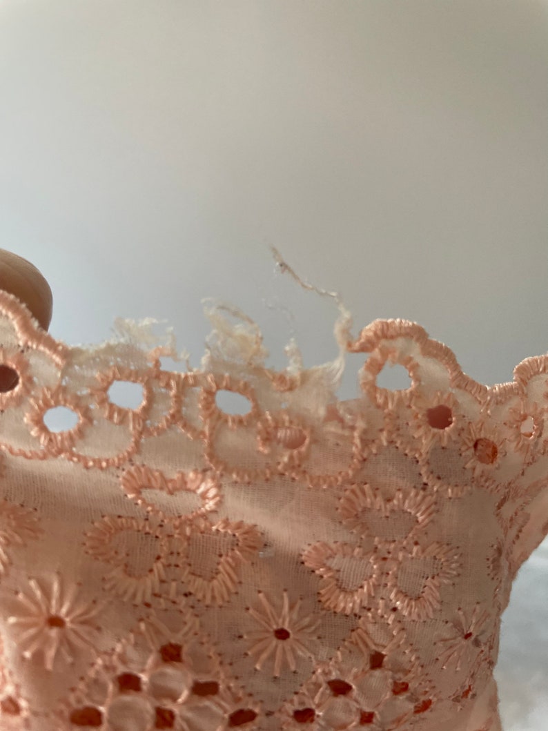 May include: A close-up of a pale pink fabric with a delicate embroidered floral pattern. The fabric has a slightly distressed look, with some of the embroidery threads frayed.