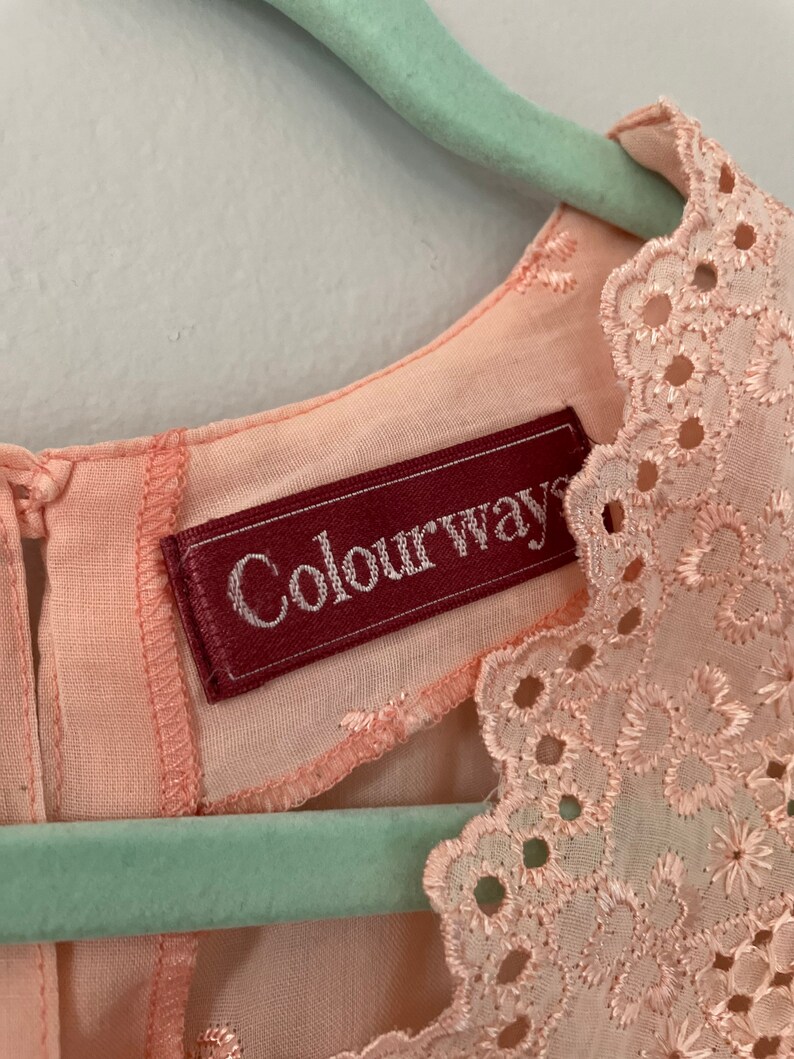 May include: A pale pink dress with a delicate lace trim around the neckline. The dress has a label that reads "Colourways".