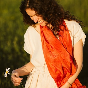 Op de afbeelding: Een vrouw draagt een crèmekleurige jurk en een feloranje sjaal. Ze houdt een madeliefje vast en glimlacht. De achtergrond is een wazig groen veld, wat een natuurlijke omgeving suggereert.