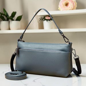 May include: A grey leather handbag with a shoulder strap and a detachable crossbody strap. The bag has a zippered main compartment and a front pocket. The hardware is silver-toned. The bag is approximately 25 cm long.