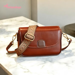 May include: A brown leather crossbody bag with a patterned strap. The bag has a rectangular shape with a curved flap and a small, rectangular accent on the front. The strap is a woven design in shades of brown, tan, and green.