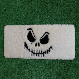Peut inclure: Un tapis de bain rectangulaire blanc avec un motif de visage de crâne noir stylisé. Le visage présente de grands yeux anguleux et un large sourire denté. Le tapis est fait d'une matière douce et moelleuse, idéal pour une salle de bain sur le thème d'Halloween.