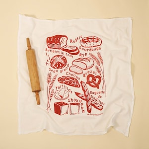 May include: A white kitchen towel with a red print of various types of bread, including sourdough, challah, tortillas, and baguettes. The text "Rustic Sourdough Rye" is printed on the towel.