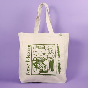 May include: A white canvas tote bag with a green graphic design featuring a person operating a sifting machine. The graphic also includes the text "New Mexico" and "Red or Green?"  The bag has a green "Eco Bag" label.