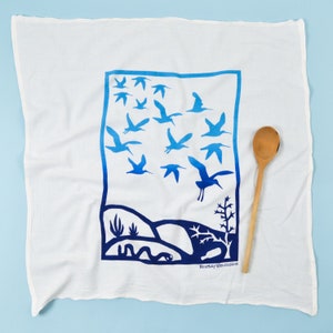 May include: White tea towel with a blue border and a design of blue birds flying over a landscape with a snake. The text "The Manly Brothers" is printed in the bottom right corner.