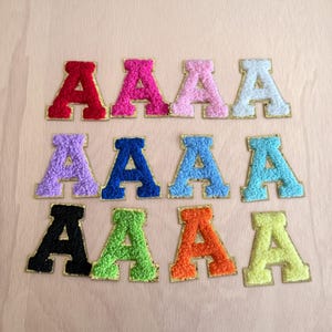 May include: Eleven embroidered letter "A" patches in various colours, including red, pink, purple, blue, black, green, orange, and yellow. Each letter has a gold outline and a textured, fuzzy appearance. Ideal for customising clothing or accessories.