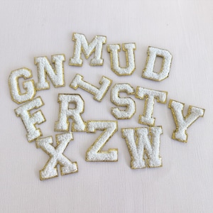 May include: White, fluffy, embroidered fabric letters with gold trim. The letters are in a variety of fonts and sizes. The letters are scattered on a light pink surface. The letters include: G, N, F, R, X, Z, W, M, U, D, S, T, Y, and V.