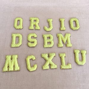 May include: A collection of fuzzy, lime green fabric letters, including Q, R, J, I, O, D, S, B, M, A, C, X, L, and U. Each letter has a gold border and is arranged on a light pink surface.