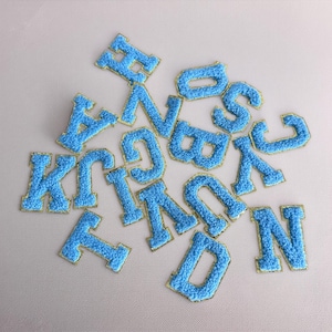 May include: A selection of blue, fluffy, embroidered alphabet letters with gold edging. The letters are arranged on a light-coloured surface. These iron-on patches can be used for customising clothing, bags, or other fabric items.