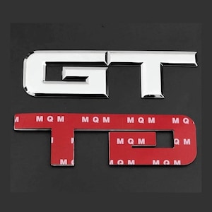May include: A pair of chrome GT car badges, one complete and one with a red adhesive backing. The letters are in a bold, reflective font. The backing has the letters "MQM" printed on it.