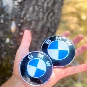 Könnte beinhalten: Zwei runde BMW-Embleme mit dem BMW-Logo, einem blau-weißen Propeller-Design, werden in einer Hand gehalten. Die Embleme sind schwarz und silberfarben, mit den Buchstaben "B", "M" und "W" um das Logo.