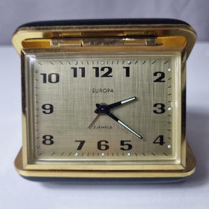 May include: A vintage gold-toned Europa travel alarm clock with a black leatherette case. The clock face features black numerals and hands, with the word "EUROPA" printed above the center. The clock is rectangular and has a hinged cover.