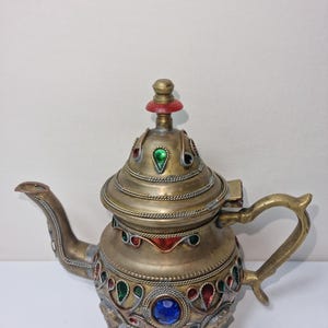 Könnte beinhalten: Verzierte Teekanne aus Messing mit dekorativem Design. Die Teekanne hat einen gebogenen Ausguss, einen Griff und einen Deckel mit roter Spitze. Sie ist mit farbigen Edelsteinen in Rot, Grün und Blau verziert und steht auf drei kleinen Füßen.
