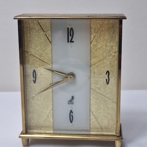 Puede incluir: Un reloj rectangular vintage en tonos dorados con una esfera de color claro. El reloj presenta números negros en las posiciones 12, 3, 6 y 9, con manecillas doradas. El reloj tiene la palabra "JAZ" y un logotipo de pájaro en la esfera.