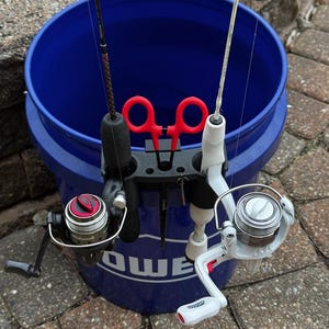 May include: A blue bucket holds two fishing rods with reels. One rod is black with a red and black reel, the other is white with a silver and red reel. A black rod holder with red accents keeps the rods in place.
