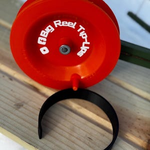 May include: Bright red fishing reel with the text "ABig Reel Tip-Ups" in white. A black, curved component sits below the reel. The reel is on a wooden surface.