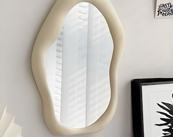 Peut inclure: Miroir mural de forme irrégulière avec un cadre crème et une section supérieure vert pâle. Le miroir est suspendu à un mur par une boucle métallique. Design moderne et artistique.