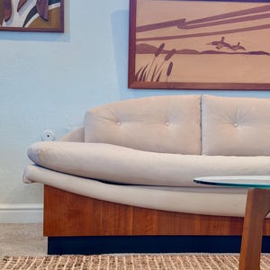 May include: A mid-century modern style sofa with a light beige upholstered seat and back, and a wooden base. The sofa is paired with a glass-topped table and a woven rug. Artwork hangs on the wall.