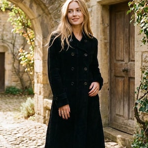 May include: A long, black double-breasted coat with large buttons. The coat is worn outdoors in front of a stone building with a wooden door and an arched entryway. The coat has a classic, tailored design.