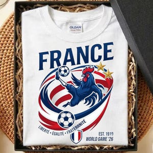 May include: A white t-shirt featuring a blue rooster kicking a football, surrounded by red, white, and blue swooshes. The text reads FRANCE, LIBERT&Eacute; &Eacute;GALIT&Eacute; FRATERNIT&Eacute;, EST. 1919, and WORLD GAME '26, with two gold stars above the rooster graphic.