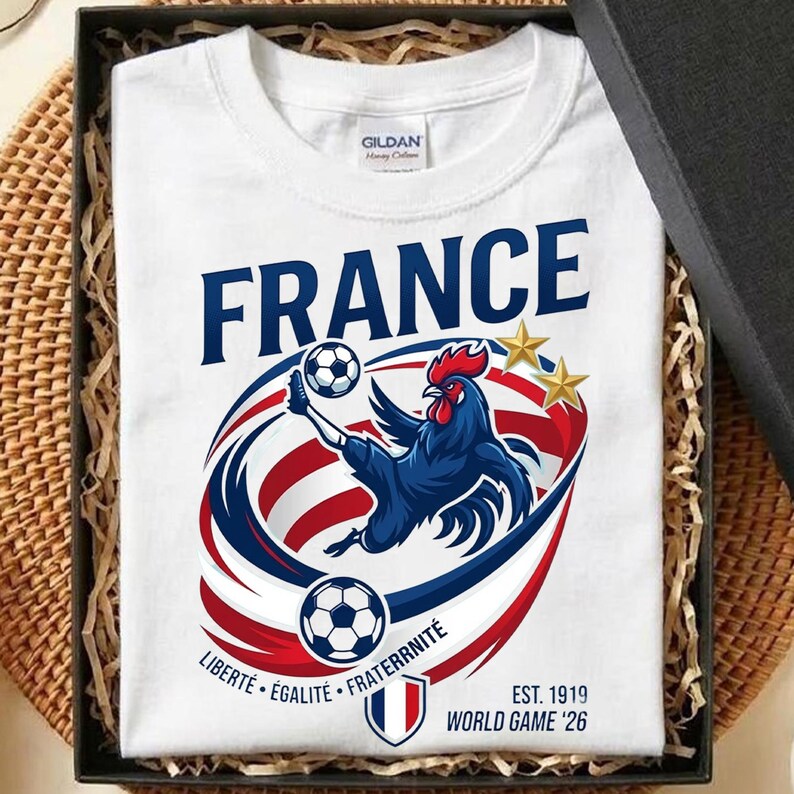 May include: A white t-shirt featuring a blue rooster kicking a football, surrounded by red, white, and blue swooshes. The text reads FRANCE, LIBERT&Eacute; &Eacute;GALIT&Eacute; FRATERNIT&Eacute;, EST. 1919, and WORLD GAME '26, with two gold stars above the rooster graphic.