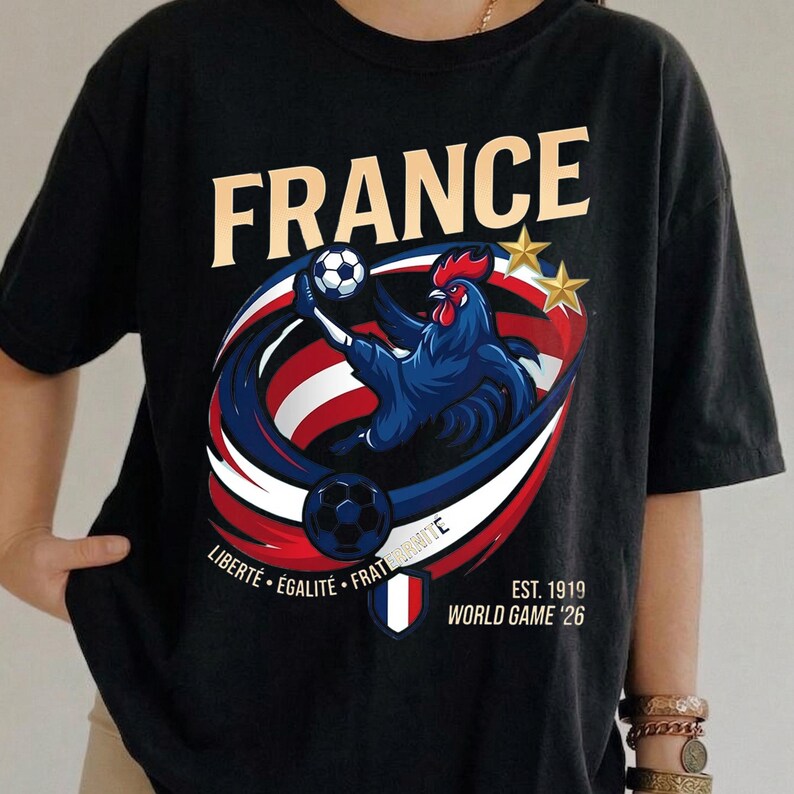 May include: A black t-shirt featuring the word FRANCE in large beige letters. The graphic shows a blue rooster kicking a football surrounded by red, white, and blue swirls, two gold stars, a football, a French flag shield, and printed text.