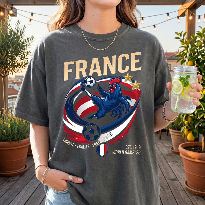 May include: A charcoal grey t-shirt featuring a graphic of a blue rooster kicking a football, surrounded by red, white, and blue swooshes. The text reads FRANCE, LIBERT&Eacute; &Eacute;GALIT&Eacute; FRATERNIT&Eacute;, and EST. 1919 WORLD GAME '26, with two gold stars.