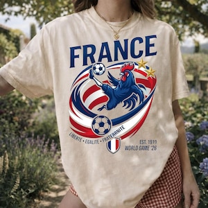 May include: A beige short-sleeve t-shirt featuring a graphic of a blue rooster kicking a football. The text reads FRANCE, LIBERT&Eacute; &Eacute;GALIT&Eacute; FRATERNIT&Eacute;, EST. 1919, and WORLD GAME '26. The design includes red and white curved stripes and two gold stars.