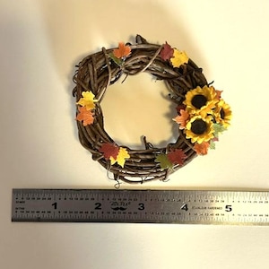 May include: A small circular wreath made of brown twisted twigs, decorated with miniature yellow, orange, and red autumn leaves and three yellow sunflowers. A metal ruler placed below shows the wreath is approximately 4 inches wide.