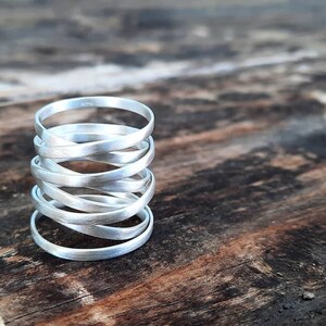 Könnte beinhalten: Ein Stapel silberner Ringe, jeder mit einer leicht unterschiedlichen Breite, sind spiralförmig angeordnet. Die Ringe sind glatt und haben eine gebürstete Oberfläche. Die Ringe liegen auf einer Holzoberfläche.