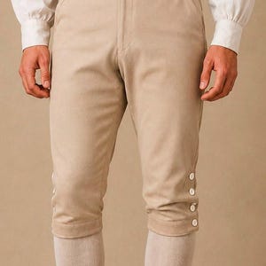 Op de afbeelding: Beige kniebroek met een hoge tailleband en drie witte knopen aan weerszijden van de sluiting aan de voorzijde. De onderkant van de broekspijpen is recht afgewerkt met vier witte knopen die verticaal aan de buitenzijde zijn geplaatst.