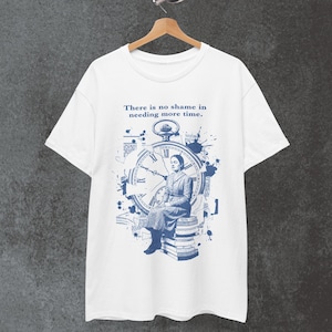 May include: White t-shirt featuring a blue graphic of a woman seated in front of a large clock face. The text "There is no shame in needing more time" is printed above the clock design. The shirt is hanging on a wooden hanger.