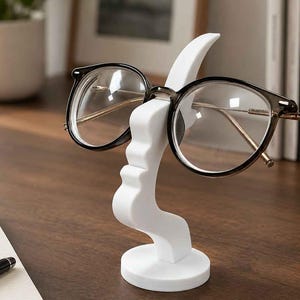 May include: A white, abstract, sculptural stand shaped like a stylised human profile, designed to hold a pair of dark-rimmed eyeglasses. The stand features a smooth, matte finish, a circular base, and a curved structure that supports the glasses.
