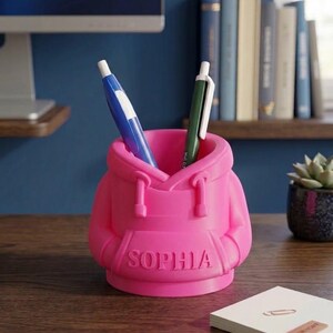 May include: A bright pink desk organiser shaped like a miniature hoodie sweatshirt with the name SOPHIA embossed on the front pocket. The organiser holds a blue pen and a green pen, featuring a hood, drawstrings, and long sleeves folded at the sides.