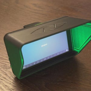 May include: A black field monitor with a green sun hood and a screen displaying the text NO INPUT. The device features a rectangular frame with a matte finish, a top-mounted logo, and a series of icons visible along the bottom of the screen.